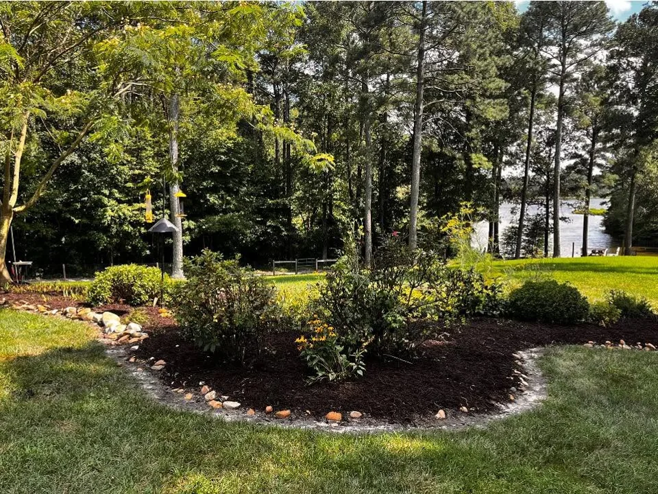 Stone-edged dark mulch and shrubs with water visible beyond the trees